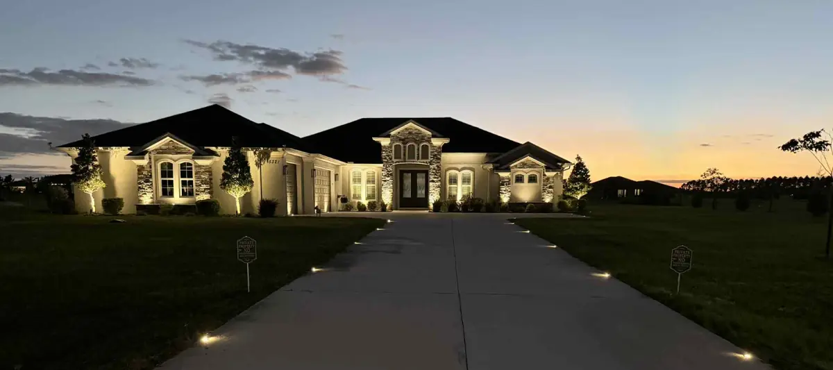 Stone home at dusk with warm up-lights on the facade and in-ground lights along the driveway, installed by Zuzack