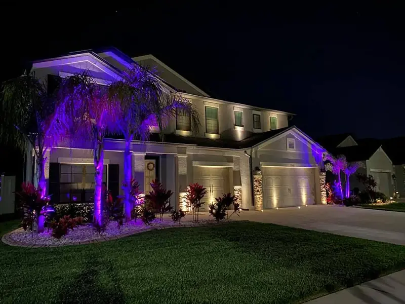 Two-story home with purple up-lighting on palms and warm light on the garage