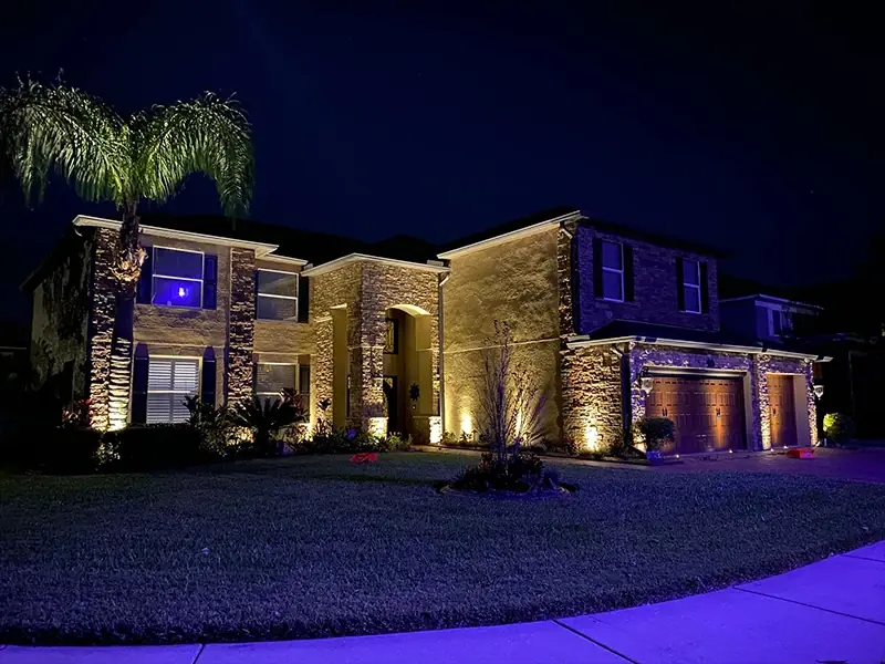 Two-story home at night with warm up-lights on stone columns, palms and the garage