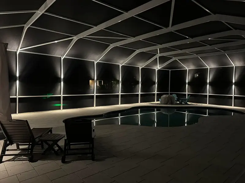 Large screened pool cage lit with warm light bars and lounge chairs by the pool