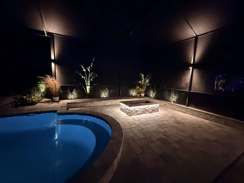 Pool and fire pit area lit warm with up-lit plants along the screen