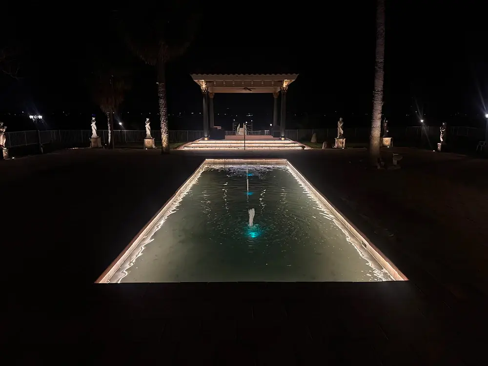 Rectangular reflection pool lit around its edge, leading to a lit pergola at night