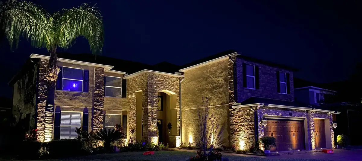 Two-story stone home lit with warm up-lights on the facade, columns and a palm at night